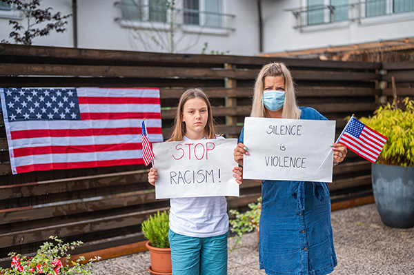 Young caucasian woman wearing a protective face mask and a little girl holding small american flags and white posters with handwritten message: STOP RACISM! and SIILENCE IS VIOLENCE. They are standing outdoors and they are looking at camera. American flag on the wooden fence in the blurred background.