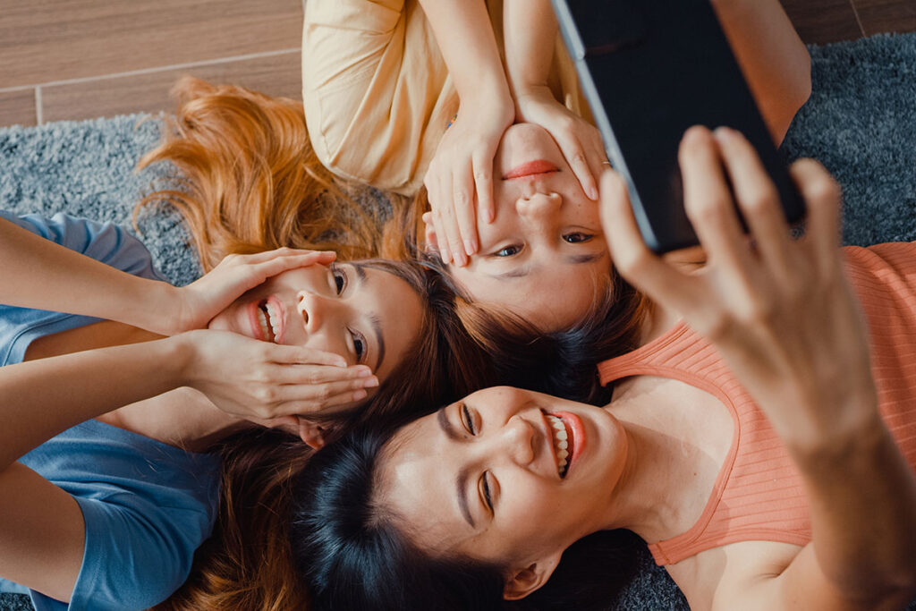 Above view close up group of Asia ladies with happiness enjoy moment hold smartphone smiling take memories picture lying on carpet in floor living room at home.
